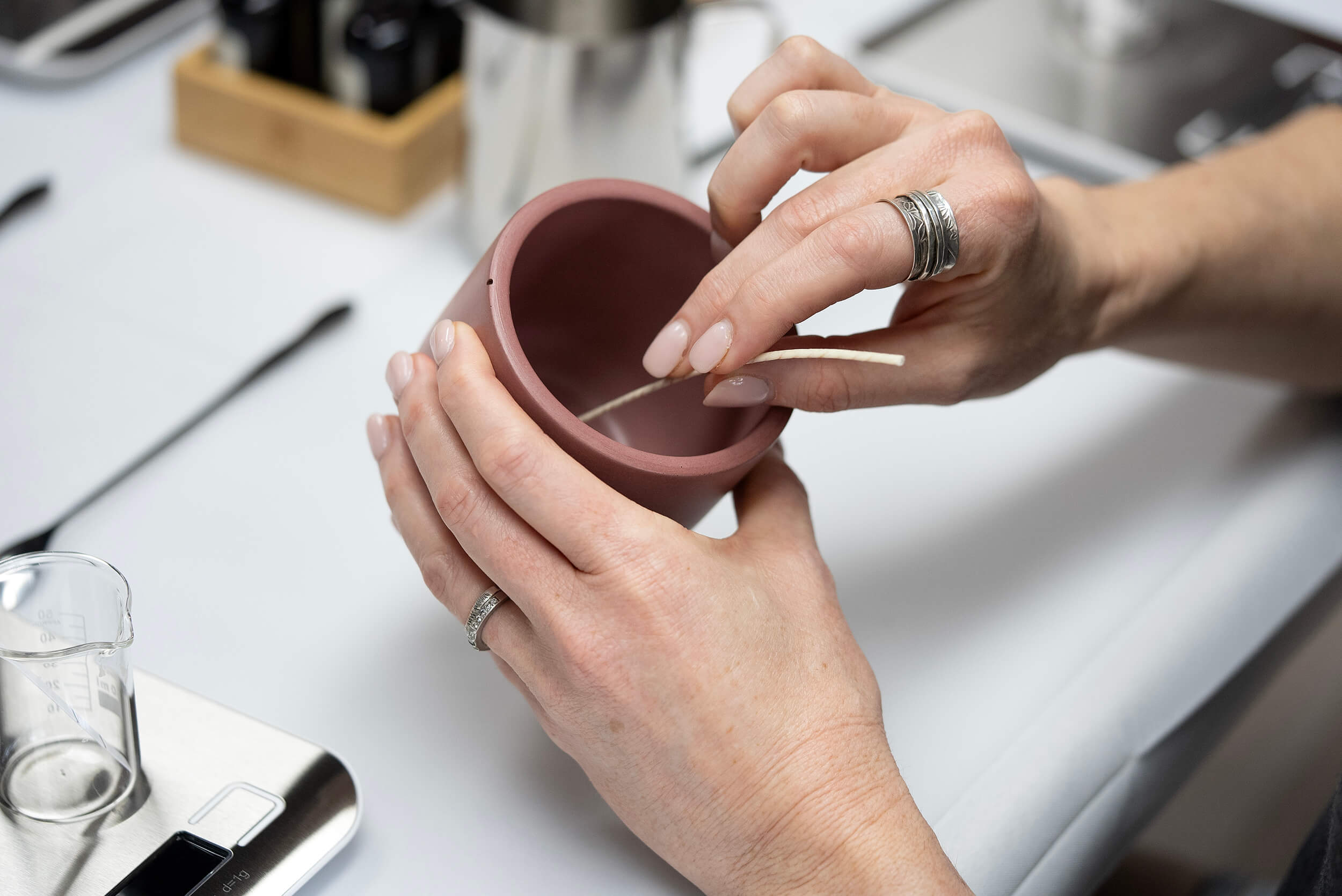 At the Candle Pouring Workshop, a participant with silver rings places a wooden wick in a clay candle holder, surrounded by a beaker and candle-making tools on the white table.