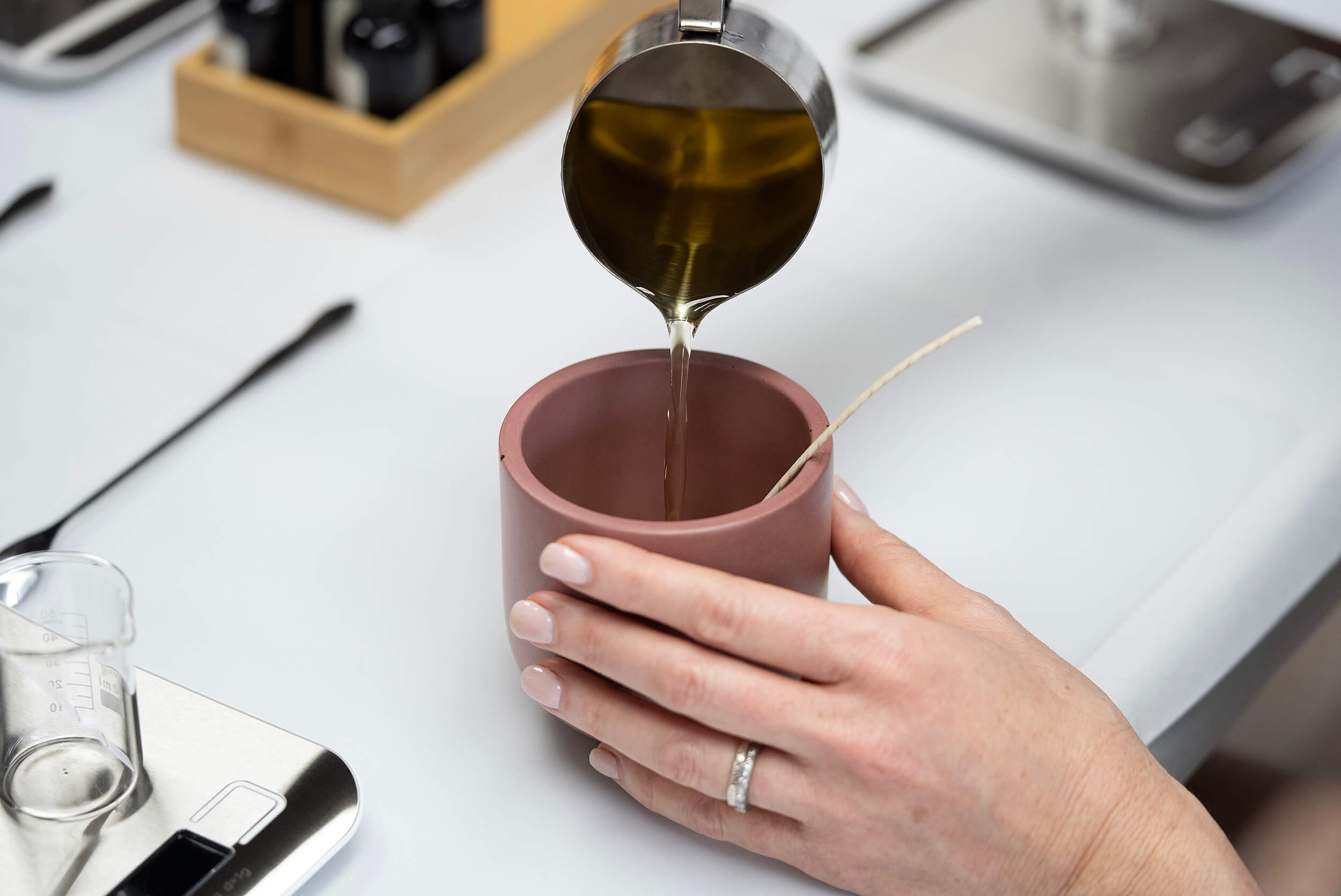 At the Candle Pouring Workshop, a person pours wax from a metal pitcher into a pink jar with a wick, steadying it on a supply-filled table to make their own custom candle.
