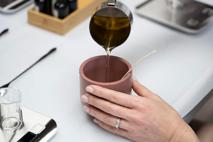 At the Candle Pouring Workshop, a person pours wax from a metal pitcher into a pink jar with a wick, steadying it on a supply-filled table to make their own custom candle.