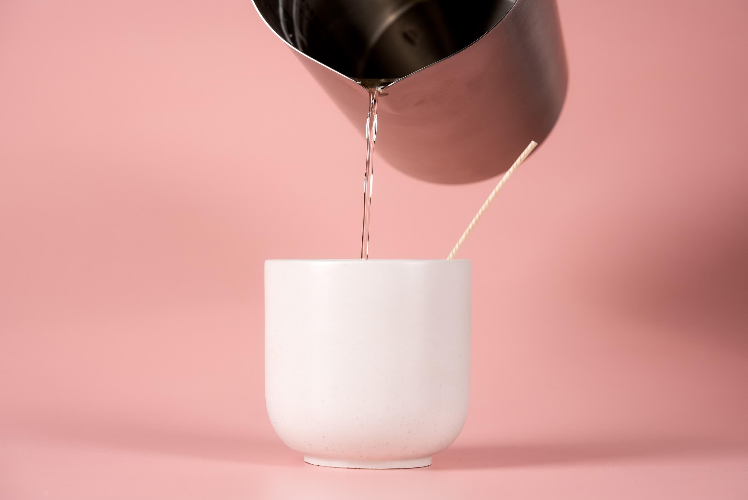 A metal pitcher pours clear liquid into a white cup with a wooden stick inside, set against a soft pink background.