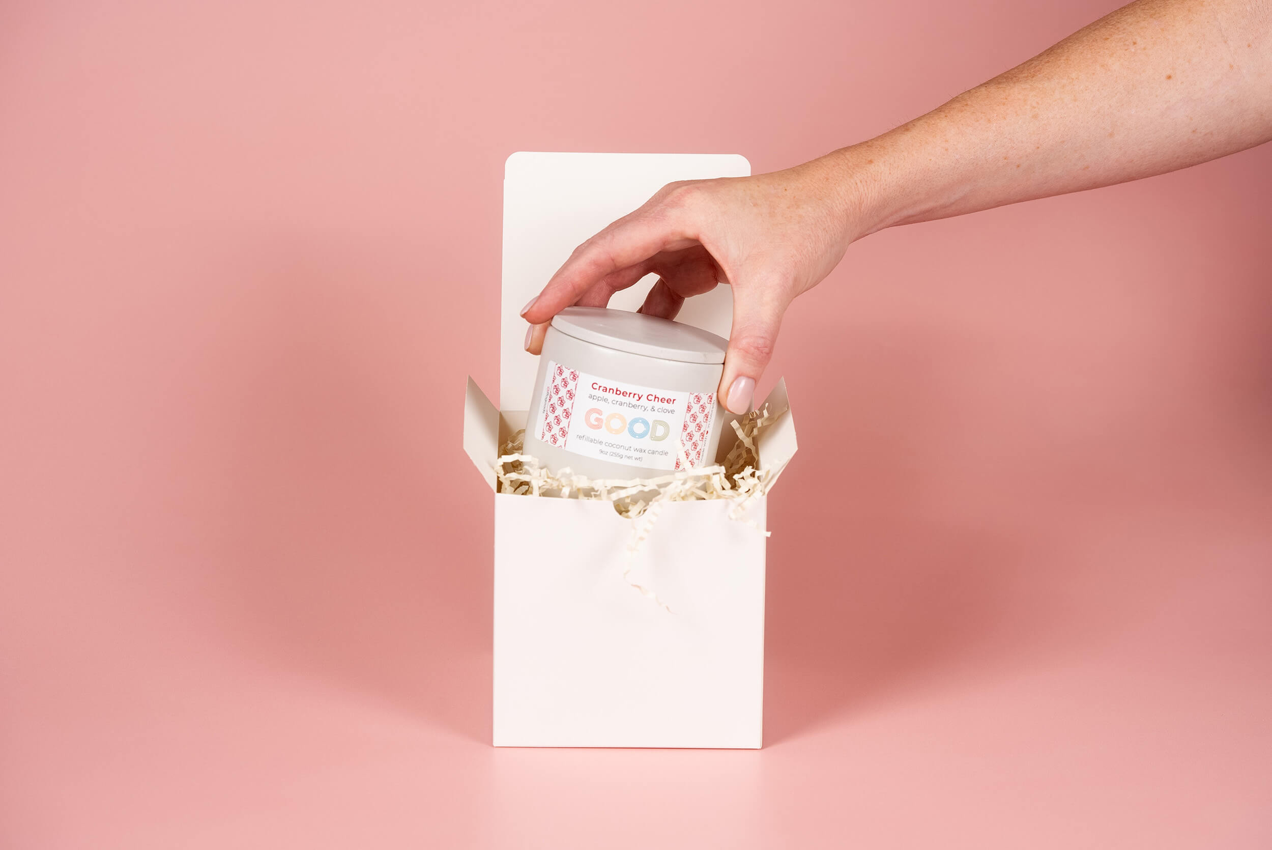 A hand places a jar labeled “Cranberry Chew” into a white gift box filled with beige paper shreds, with a pink background.