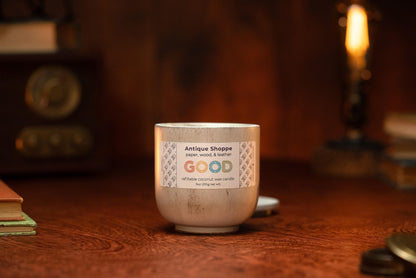 The Antique Shoppe candle in a metallic container sits on a wooden table, surrounded by books and warm ambient lighting, with a vintage radio in the background.