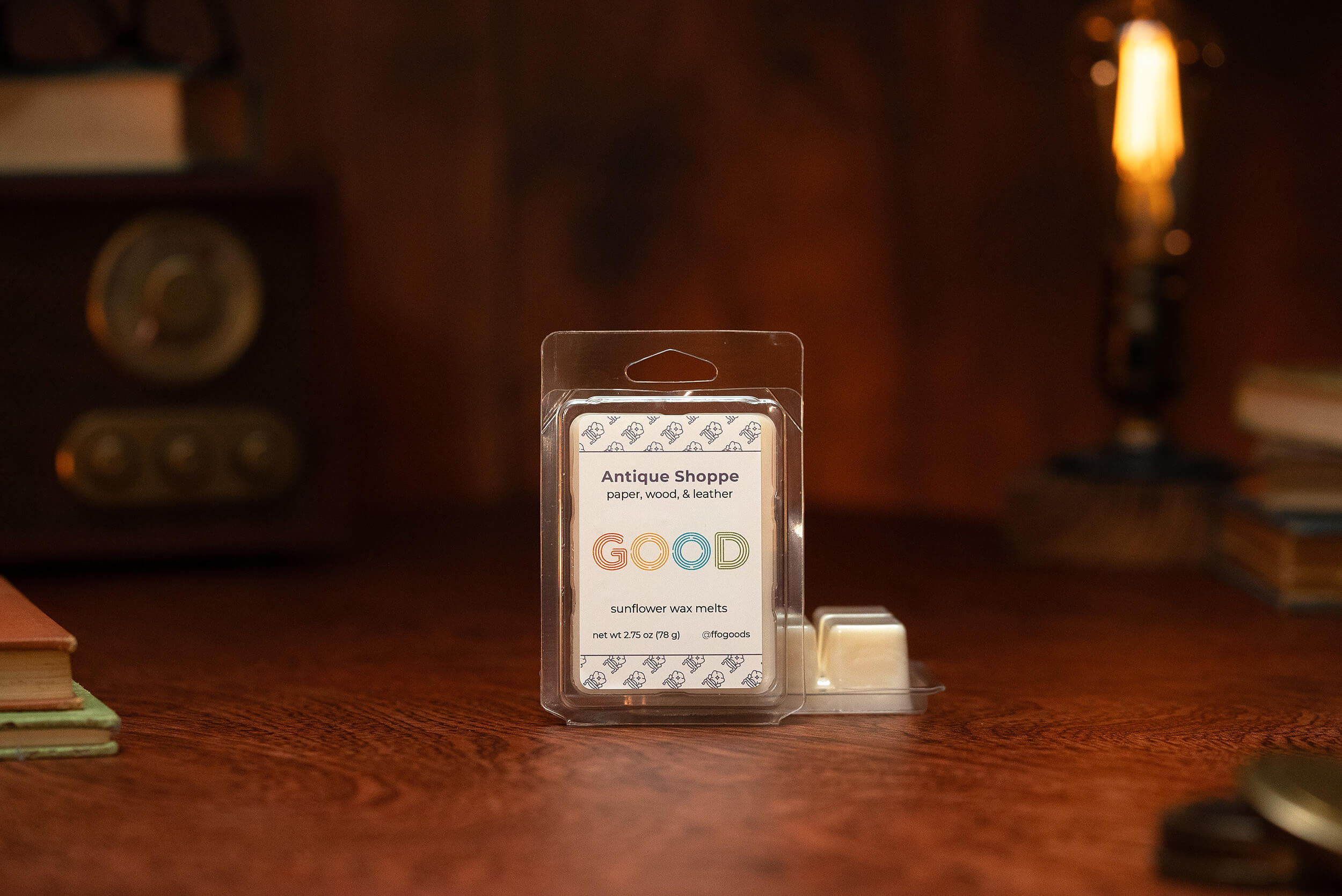 A package of Antique Shoppe Wax Melts sits on a wooden table with one melt removed. A vintage radio, books, and a glowing Edison bulb lamp in the background evoke coziness and long-lasting fragrance.