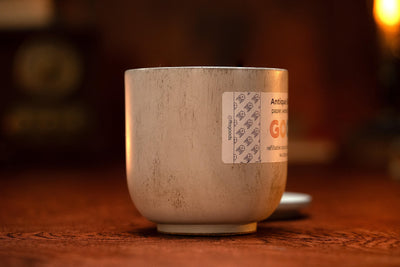 A close-up of a white ceramic cup with visible scratches, featuring the "Antique Shoppe" candle label, sits on a wooden surface against a softly lit, blurred background.