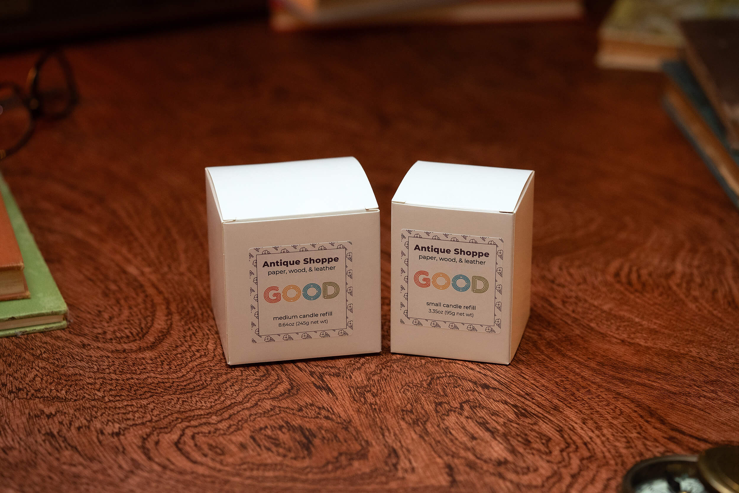 Two white Candle Refills boxes labeled Antique Shoppe with "GOOD" logo, one medium and one small, rest on wood. Both candles feature paper, wood, and leather scents in biodegradable packaging.