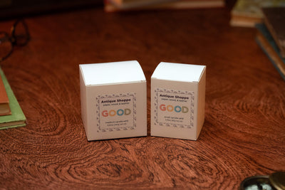 Two white Candle Refills boxes labeled Antique Shoppe with "GOOD" logo, one medium and one small, rest on wood. Both candles feature paper, wood, and leather scents in biodegradable packaging.