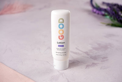 A white tube of Hydrating Body Lotion stands upright on a light purple surface, with blurred greenery and purple flowers in the background. The label displays colorful, playful lettering.