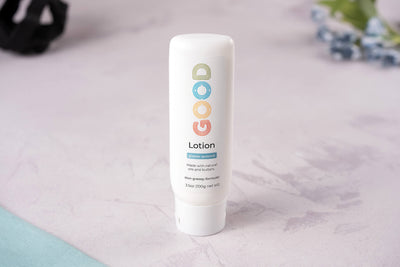 A white bottle of Hydrating Body Lotion stands upright on a light, textured surface. The label has colorful letters and highlights a glacier summer scent with nourishing ingredients. Blueberries and fabric appear blurred in the background.