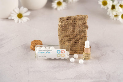 Scent Diffusing Ceramic Beads, a small glass bottle of long-lasting “Fresh Linen” fragrance beads, come with a burlap pouch and dropper bottle, all arranged on a light surface with daisies in the background.