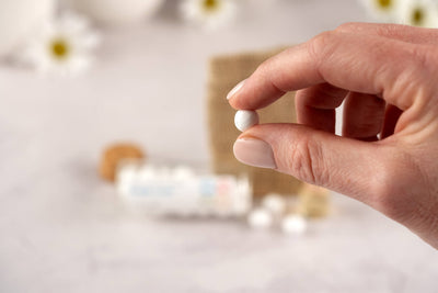 A close-up of a hand holding a small white round Scent Diffusing Ceramic Bead between the thumb and index finger, with a blurred background showing more beads, a bottle, flowers, and additional eco-friendly air freshener products.