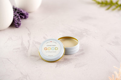A small white tin of Cuticle Cream with the lid off reveals the cream inside. Made with natural ingredients, it sits on a light pink textured surface, with lavender and greenery in the background.