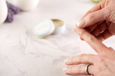 A person applies Cuticle Cream to their finger from an open tin on a light marble-like surface, focusing on attentive nail care with the product tin softly blurred in the background.