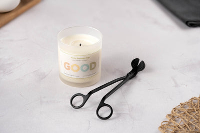 A white candle in a glass jar labeled GOOD sits on a light surface next to the Wick Trimmer, the perfect accessory for thoughtful candle care. A small piece of woven material appears in the bottom right corner.