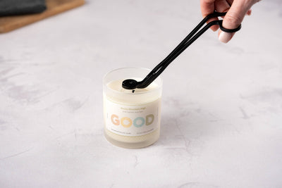 A hand uses the Wick Trimmer to neatly trim the wick of a white candle in a glass GOOD jar, demonstrating essential candle care on a light surface.