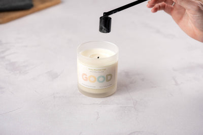A hand uses a Candle Snuffer to extinguish a white candle in a glass jar labeled "GOOD" on a light surface, with smoke rising from the candle.
