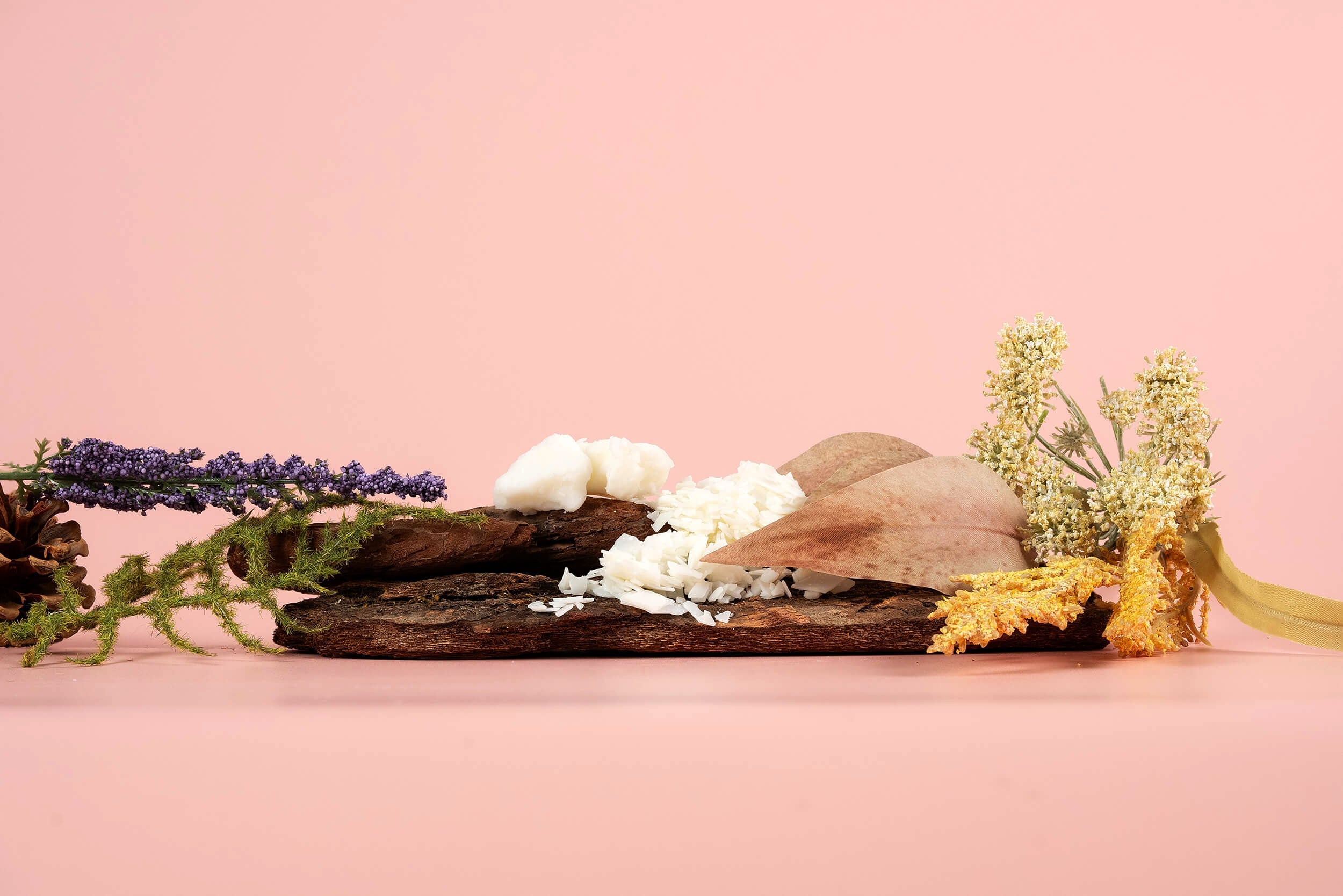 A piece of bark holds white flakes, a chunk of white substance, brown leaves, and is surrounded by sprigs of purple, yellow, and green flowers against a pink background.
