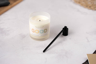 A white candle in a glass jar labeled GOOD with Rocky Mountain High scent—pine, sage, and lavender—rests on a light surface beside the Candle Snuffer, a sleek black accessory for safely extinguishing candles.