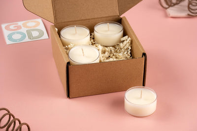 A cardboard box from FFO Goods contains four Sample Sniffs tea-light candles nestled within shredded paper. One candle is placed outside the box on a pink surface, while a card displaying the word GOOD in colorful letters suggests a delightful scent variety.