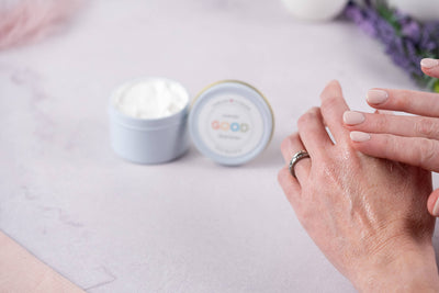 A person applies Whipped Body Butter to the back of their hand near an open container of white cream on a light surface. They have neatly manicured nails and wear a silver ring.