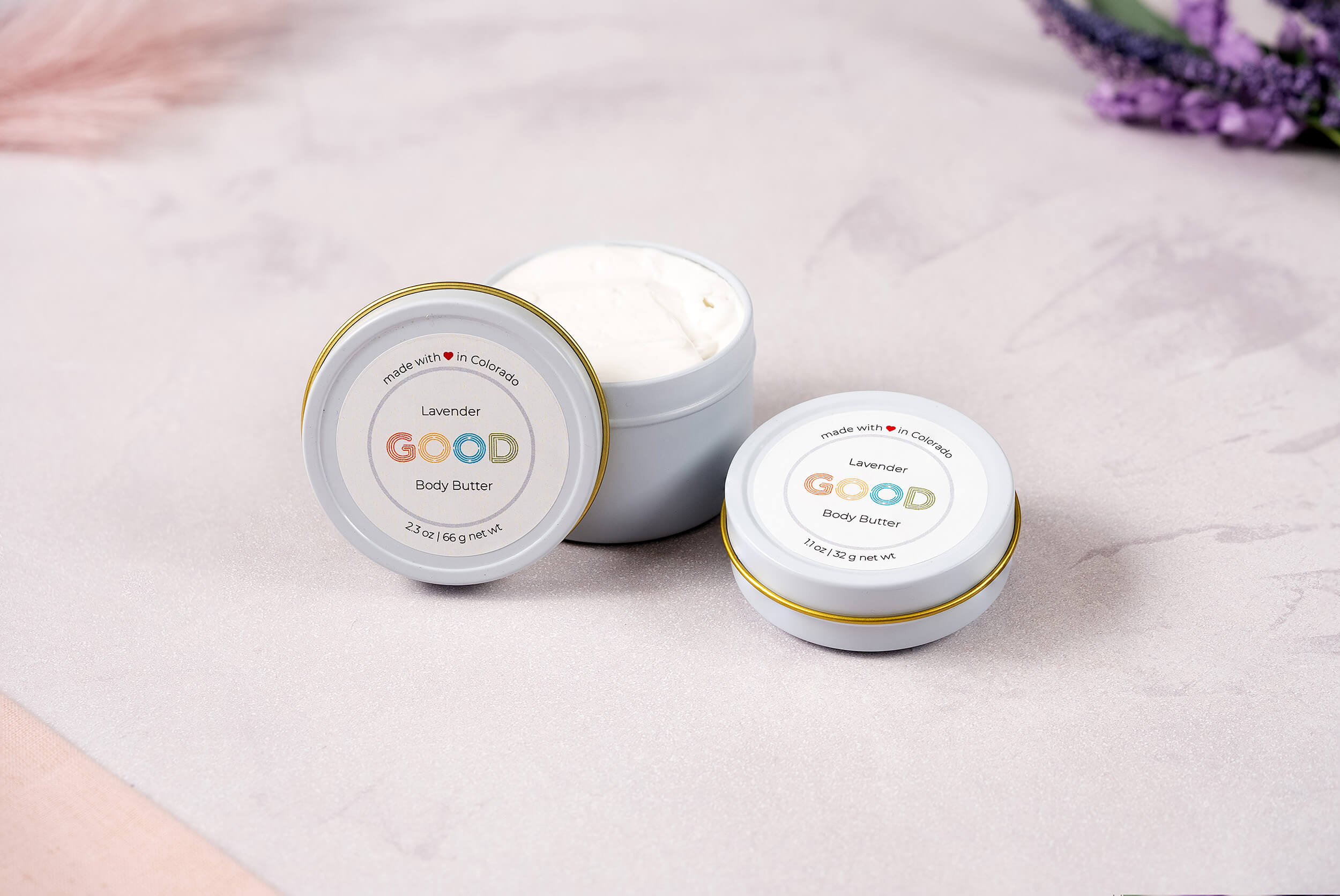 Three small white tins of Whipped Body Butter sit on a soft pink and white surface. Two tins are closed, showing the lavender label; one is open, displaying the creamy vegan formula inside.