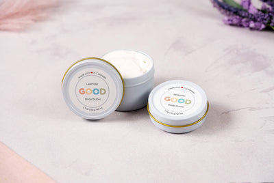 Three small white tins of Whipped Body Butter sit on a soft pink and white surface. Two tins are closed, showing the lavender label; one is open, displaying the creamy vegan formula inside.