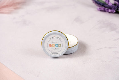 An open tin of Whipped Body Butter labeled GOOD rests on a light, textured surface with soft pink and purple accents in the background.