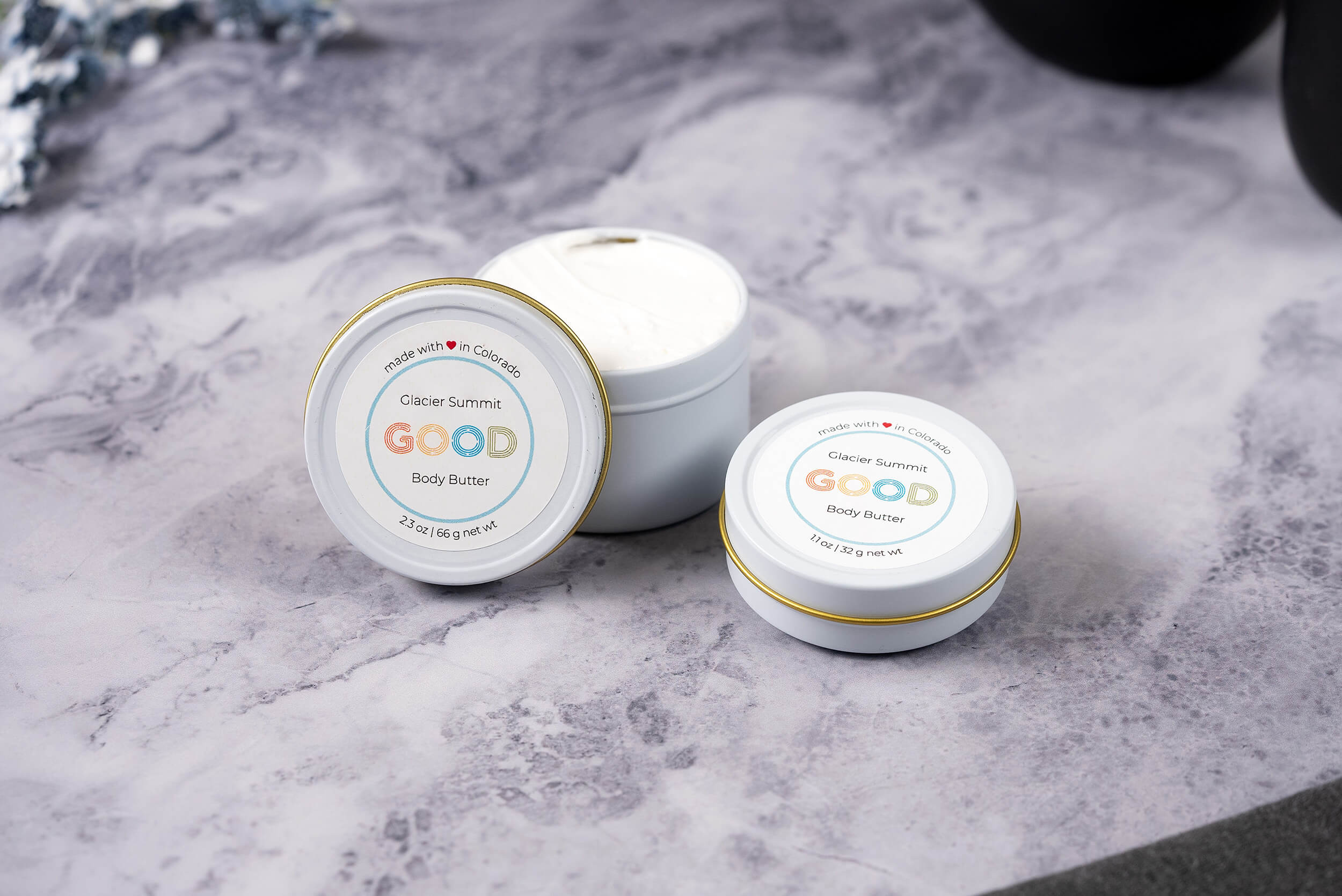 Two tins of Whipped Body Butter, prized for ultra-moisturizing vegan care, rest on a gray marbled surface—one open to show the creamy texture. Each has a white lid with a simple, colorful label.