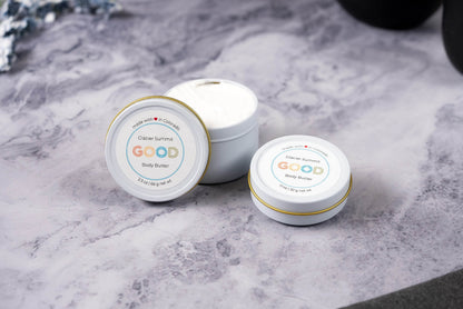 Two tins of Whipped Body Butter, prized for ultra-moisturizing vegan care, rest on a gray marbled surface—one open to show the creamy texture. Each has a white lid with a simple, colorful label.