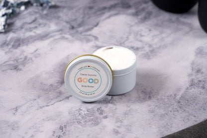 A small white jar of Whipped Body Butter sits open on a marbled surface, revealing rich cream inside. The visible lid label highlights its vegan skincare benefits and ultra-moisturizing formula.