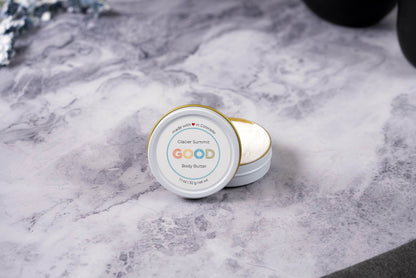 An open tin of Whipped Body Butter rests on marble, revealing its creamy texture inside. The white lid leans against the tin, showcasing this ultra-moisturizing skincare treat.
