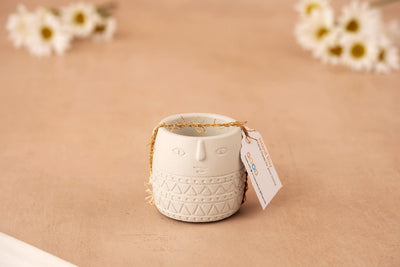 The Island Boy – Concrete Character Vessel is a small white air plant holder with a face design, gold string, and tag. Set on a soft beige surface with blurred white daisies, it's perfect minimalist desk decor.
