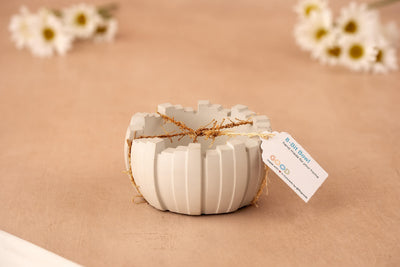 The 8 Bit Bowl – Retro Meets Modern is a round, white geometric concrete bowl tied with straw and a tag, displayed on a beige surface with blurred white daisies in the background.