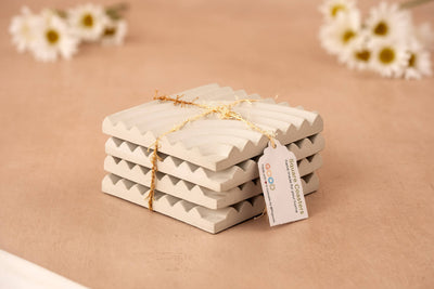 A set of four white Concrete Coasters, tied with gold string and labeled “Concrete Coasters – Set of 4,” rests on a beige surface. Blurred white daisies in the background highlight this eco-friendly coaster set.