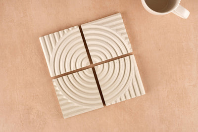 The Concrete Coasters – Set of 4 features white geometric coasters with circular grooves, arranged to create a unified design on a light beige surface. A white mug is placed in the upper right corner for a modern touch.