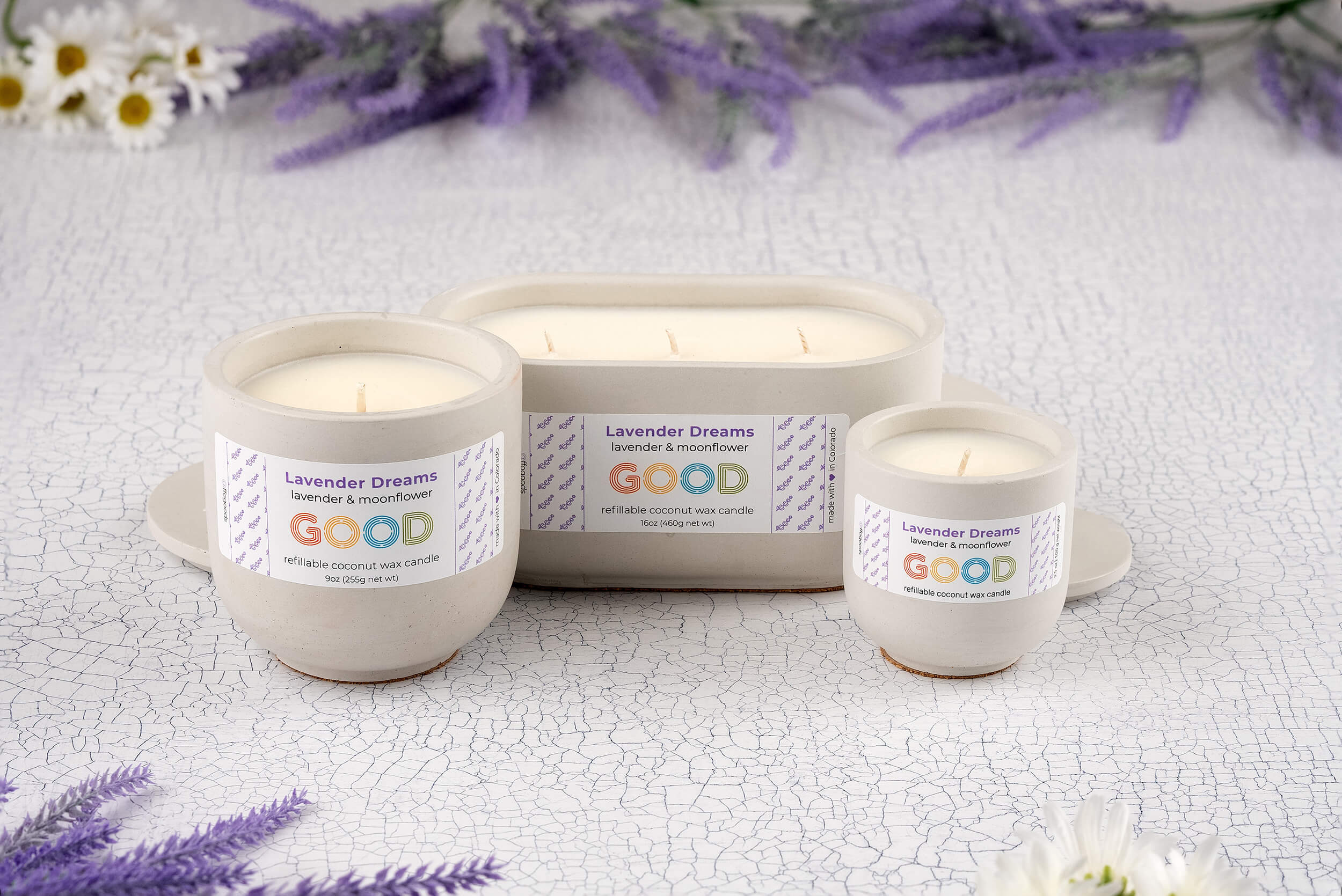 Three Lavender Dreams candles in hand-poured white concrete vessels are displayed on a cracked white surface, surrounded by purple lavender and white flowers. These sustainable candles come in simple, elegant containers.