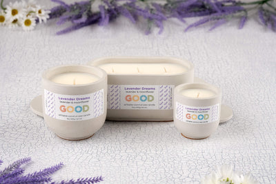 Three Lavender Dreams candles in hand-poured white concrete vessels are displayed on a cracked white surface, surrounded by purple lavender and white flowers. These sustainable candles come in simple, elegant containers.