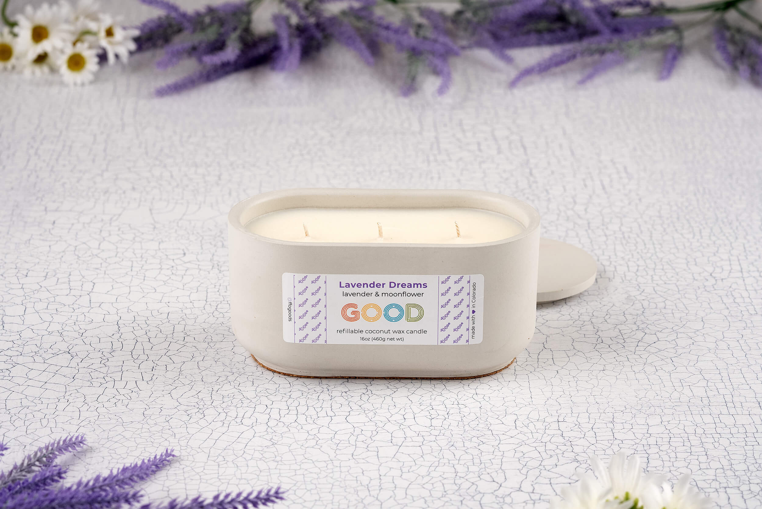 The Lavender Dreams candle, a white oval in a hand-poured concrete vessel, rests on a textured white surface surrounded by purple and white flowers, its lid set aside.