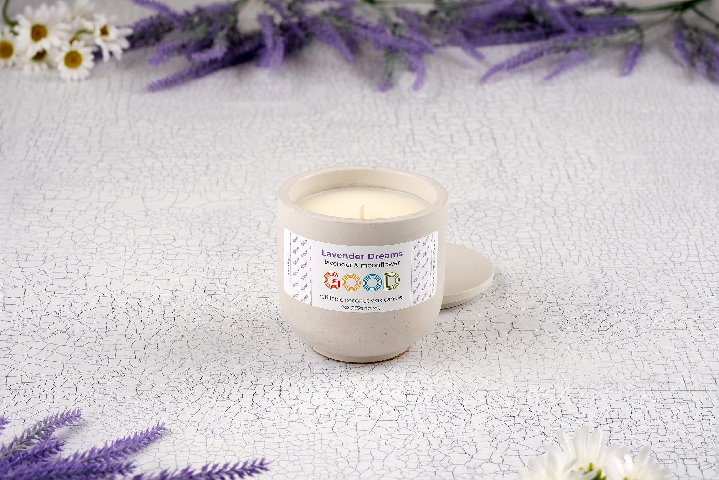 A hand-poured Lavender Dreams candle sits on a textured white surface, surrounded by purple lavender sprigs and small white flowers. Its label reads "lavender + moonflower" and "GOOD.