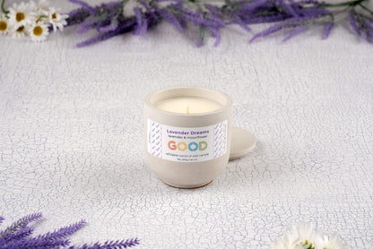 A hand-poured Lavender Dreams candle sits on a textured white surface, surrounded by purple lavender sprigs and small white flowers. Its label reads "lavender + moonflower" and "GOOD.