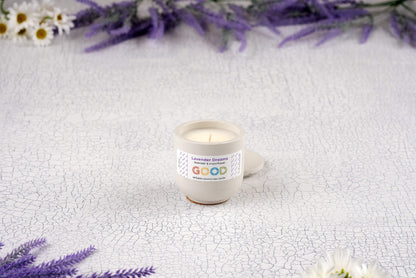 A Lavender Dreams candle in a hand-poured concrete vessel sits on a cracked white surface, surrounded by white daisies and sprigs of purple lavender.