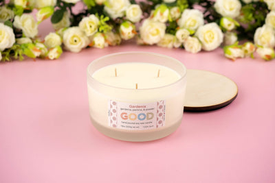 A white three-wick soy candle named Gardenia sits in a frosted glass jar with a wooden lid, placed on a pink surface and surrounded by white and cream roses. The label reads "Gardenia GOOD" with scent notes and candle details.