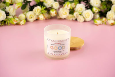 A “Gardenia” candle sits on a pink surface in a frosted glass jar with a wooden lid. The label highlights gardenia, jasmine, and powder notes. Softly blurred white flowers appear in the background.