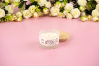 The Gardenia candle, made from natural soy wax and topped with a wooden lid, sits on a pink surface with white roses in the background, filling your space with its delicate floral scent.