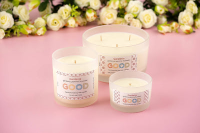 Three white Gardenia-scented natural soy wax candles in frosted glass jars of assorted sizes sit on a pink surface with white flowers behind them. Each candle features a GOOD label.