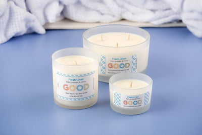 Three white soy wax candles in frosted glass jars labeled "Fresh Linen" are arranged on a blue surface, with a folded white towel in the background, evoking the clean and refreshing scent of Fresh Linen.