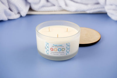 The Fresh Linen three-wick soy candle, in a frosted glass holder with a wooden lid, sits on a blue surface. Its label reads "Fresh Linen" and "GOOD," releasing clean linen, powder, and citrus scents. A white towel is in the background.