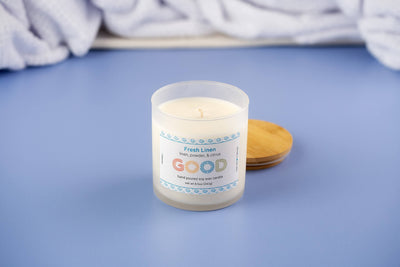 The Fresh Linen candle in a frosted glass jar with a wooden lid rests on a blue surface, releasing a clean linen scent as a white textured fabric appears softly blurred in the background.