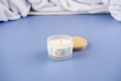 The Fresh Linen soy wax candle rests in a frosted glass jar on a blue surface, its wooden lid nearby. A soft white towel in the background hints at the crisp, clean linen scent of this product.