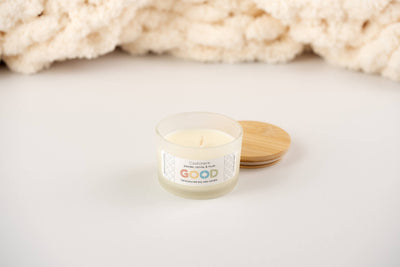 A small hand-poured soy wax candle named Cashmere, with a wooden lid, rests on a white surface against a cream-textured background. The label reads GOOD and features the luxurious scent of cashmere.
