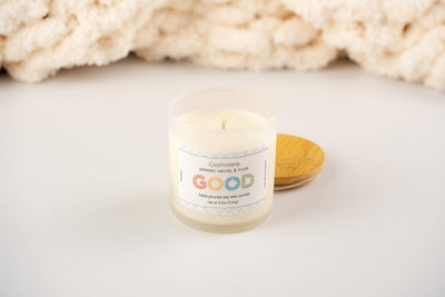 A Cashmere-scented candle labeled "GOOD" sits on a white surface beside its wooden lid, housed in frosted glass; a soft cream fabric forms the background.